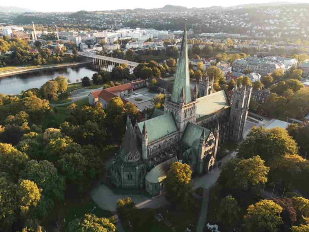 A large stone cathedral with towers and speers surrounded by big green treese and parks, with a river in the background, and lots of houses of a small town. Things to do in Trondheim. 