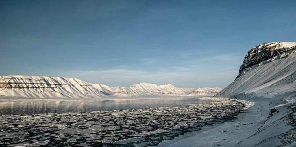 An icey fjord in an artcic landscape on a sunny day, the ice is cracking up into small ice flakes under the blue sky, surrounded by snowy mountains