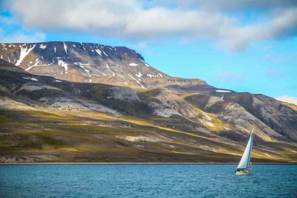 A small sailboat on the deep blue waters outside a barren landscape of grassy and rocky mountainsides with no trees, and pathces of snow at the top of the mountain on a sunny day