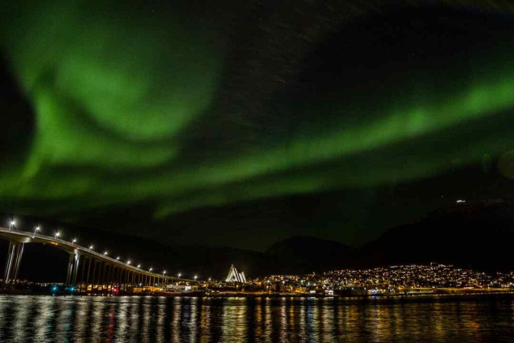 Green northern lights dancing across the black night sky over a town with thousands of lights