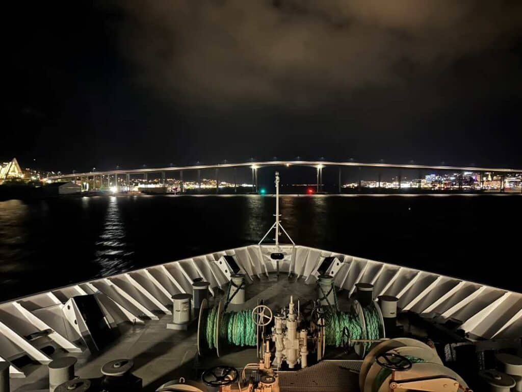 Is Trosø worth visiting? The bau of a ship at night, lit by white lights, and in front of the ship over the dark water you see a wide bridge also lit that crosses the water, where the ship will sail under