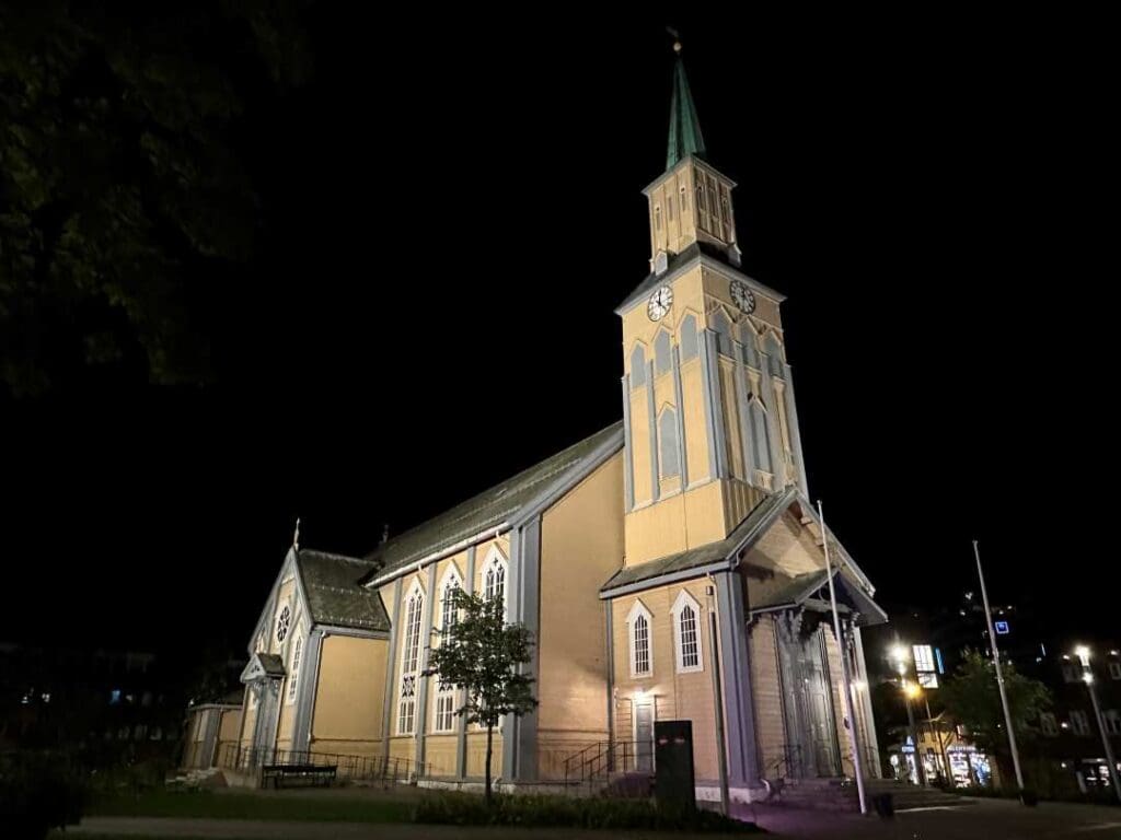 An old wooden church at night, in a pale yellow with grey decorations, a tall bell tower against the black night sky