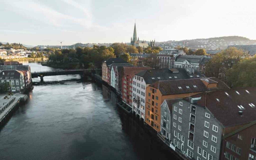 Aerial photo of a dark blue river running in the middle of colorful wooden houses, a bridge is in the distance, and the spear of a cathedral is in the distance under a pale blue sky. Best Hotels In Trondheim