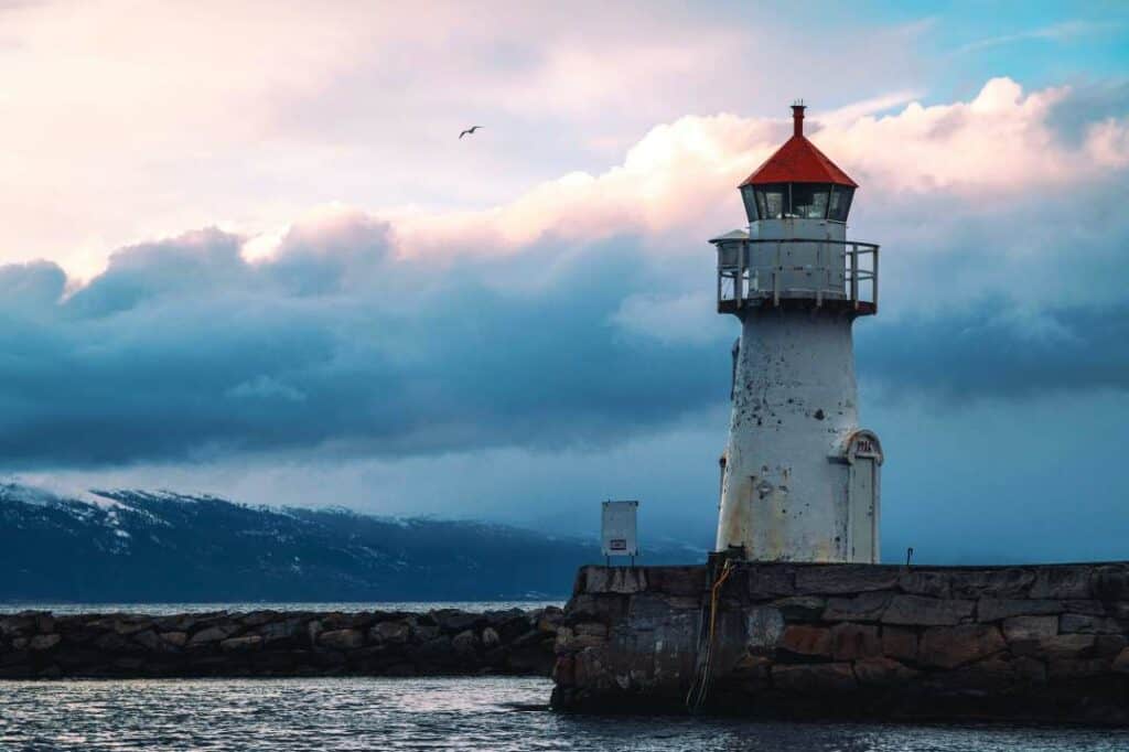 A white stone lighthouse with a red roof standing on a stone barrier from the sea with dark hills in the background across the water