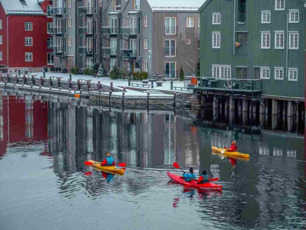 Three kayakers paddling on a dark river in winter, surrounded by old wooden houses in a small town