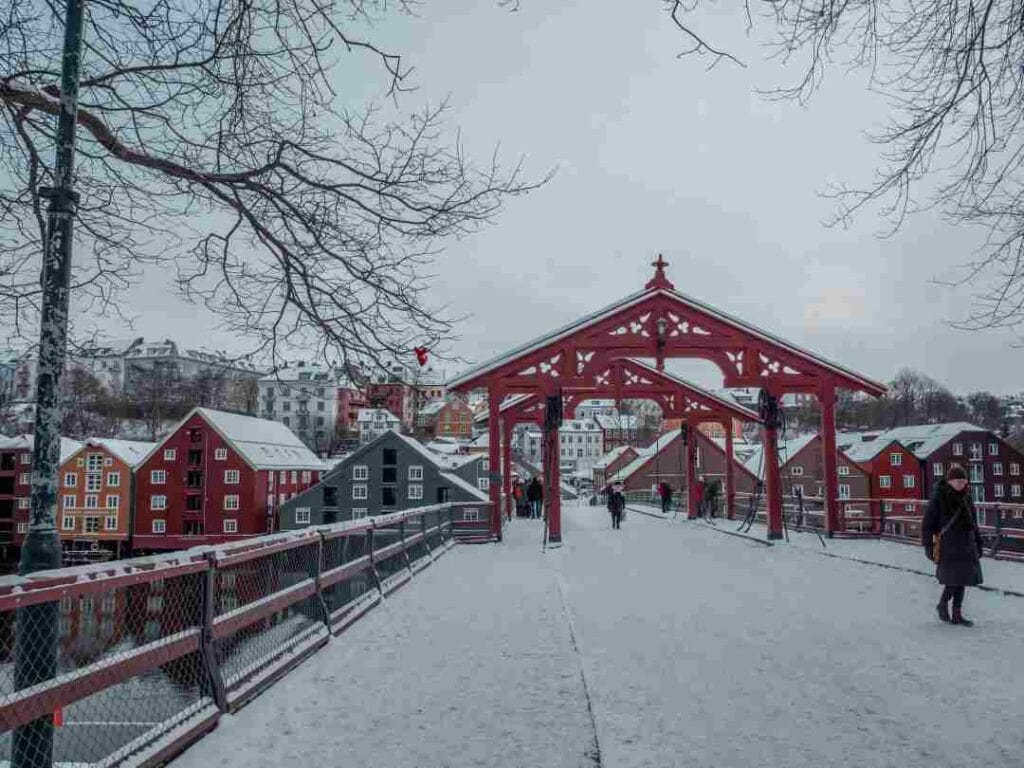 A winter bridge covered in snow on a cold day with white light, naked trees, and colorful wooden houses in the distance across the bridge