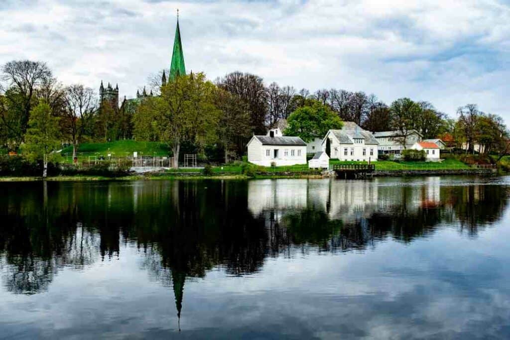 White wooden houses among green trees and parks across a water that is still and blank, under a cloud dotted sky