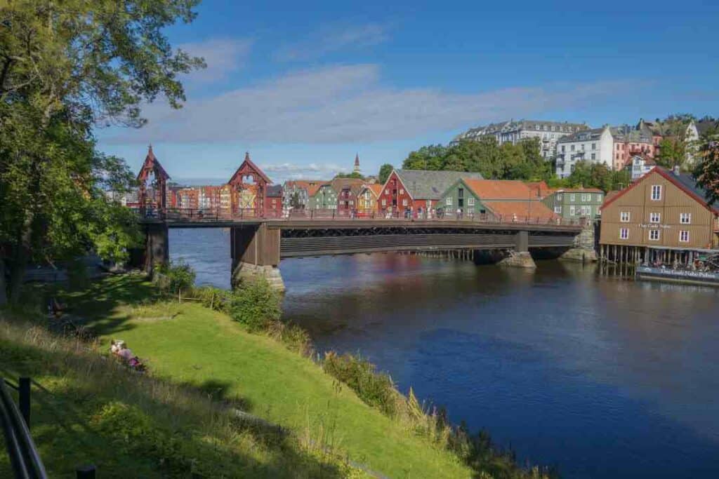 Picturesque grassy riverside area in front of a dark blue river, with a charming bridge crossing the river, and colorful wooden houses on the other side of the water under blue skies