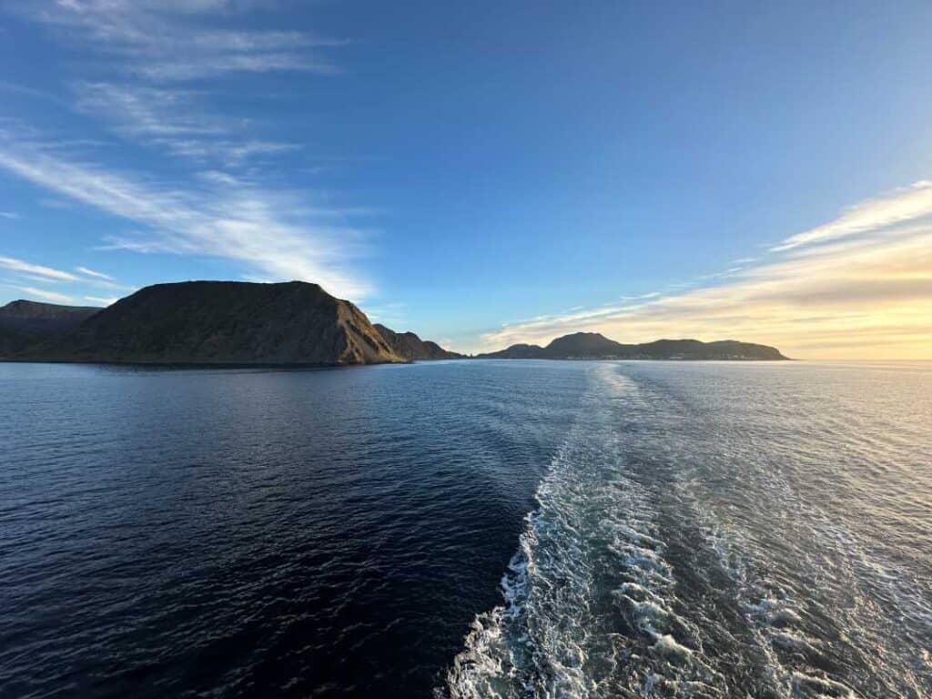 The white wake water behind a ship right before sunset under a pale blue sky above the water, with the glow of the sun from outside the photo frame