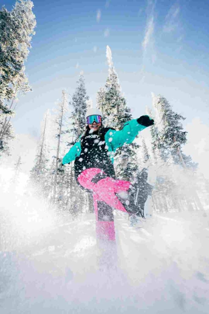 A smiling young girl in Alpine skiing outfit, with goggles, boots, and colorful ski dress in kicking dry white snow towards the photographer on a clear winter day with snow hanging on the tall trees around her