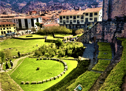 An vibrant green park with circular ruin formations in an old mountain town next to an old stone convent, with views of the mountains in the background under a blue sky