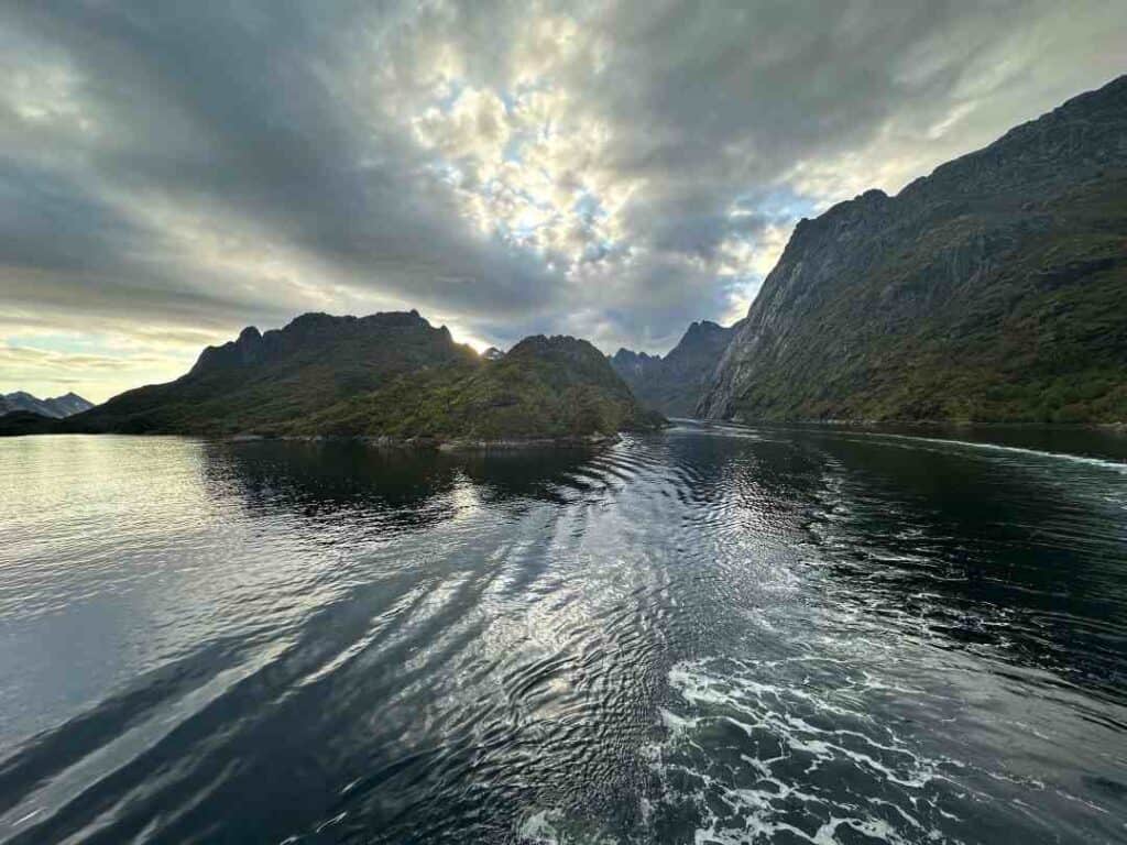 The white trail of the wake behind a ship just exiting a majestic fjord in the Arctic during summer with pale hazy light. Norwegian coastal voyage. 