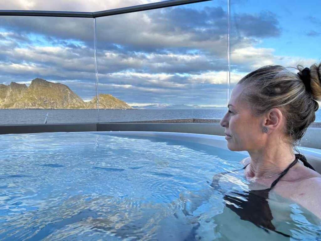 A lady sitting in a hot tub on the deck of a ship at sea, whatching stunning mountain scenery slowly passing by