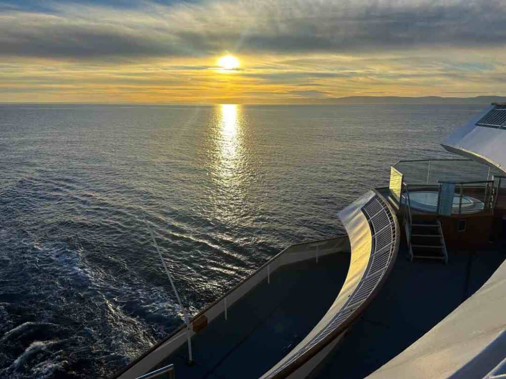 A glowing setting sun in the horizon over a dark blue ocean, seen from the bau of a ship that looks beautiful in the golden sunlight