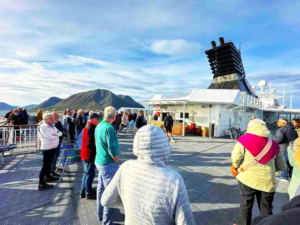 People standing on deck of a ship, dressed for cold weather with down jackets, hats and mittens, the sun is shining but it is obvioudly chilly, and there are arrid mountains in the background. 