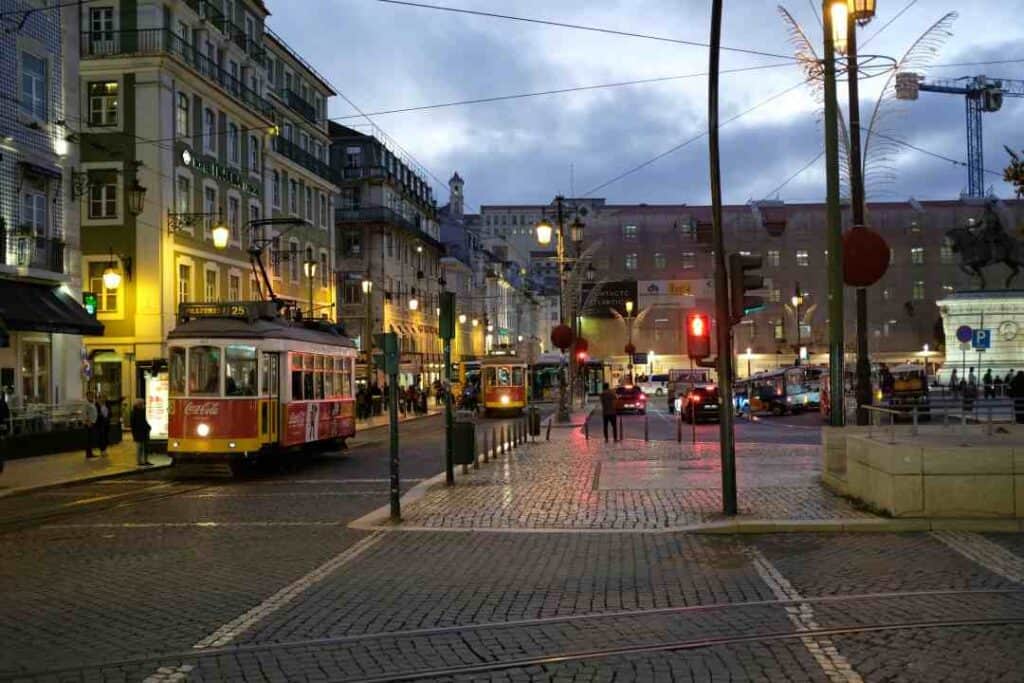 A town square at dusk with cobblestones, an old fashioned tram, lights, traffic, and people amidst the old colonial buildings lit by street lights