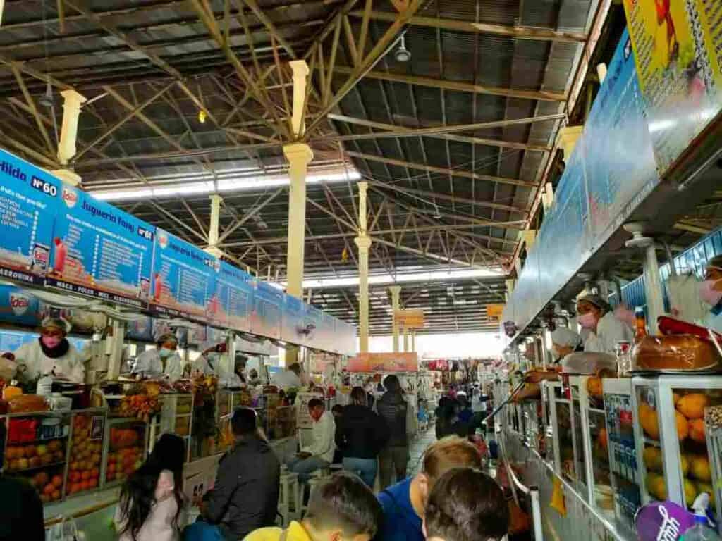 A busy market under a tall ceiling with stalls where vendors sell fruit 