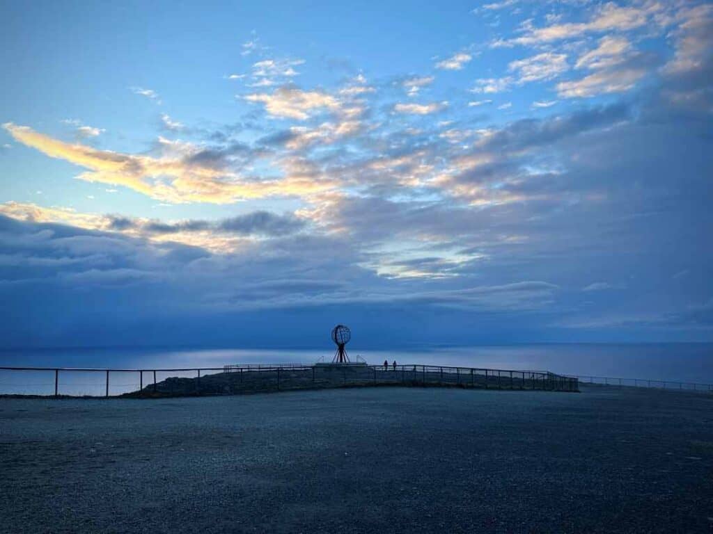 An iconic iron globe standing at the tip of a huge cliff 300 meters above the sea, amidst blue lights from the Arctic dusk, with nothing but dark Arctic water in front reaching all the way to the North Pole