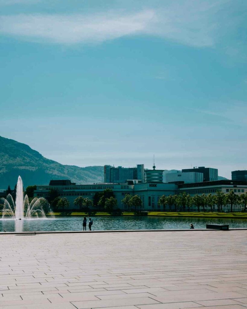 A city park with a large lake with a fountain i the middle and people strolling on the shore on a sunny day