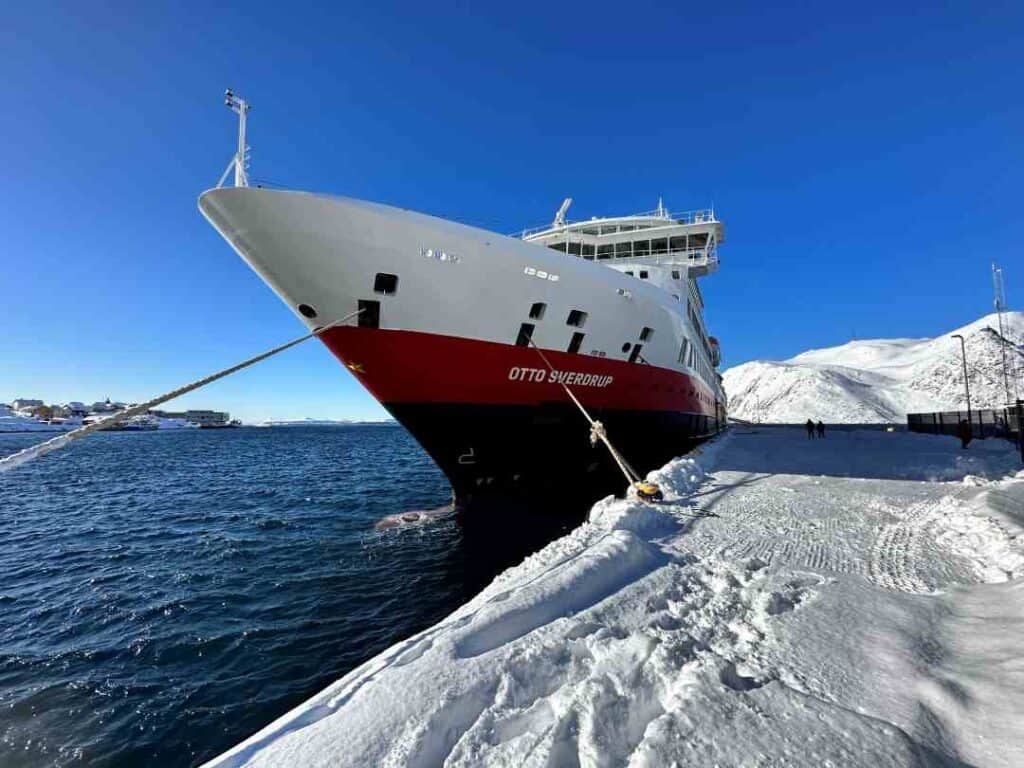 A red and white ship docked at a port covered with white snow, under a deep blue sky, with white snow covered mountains in the distance across the dark blue choppy water