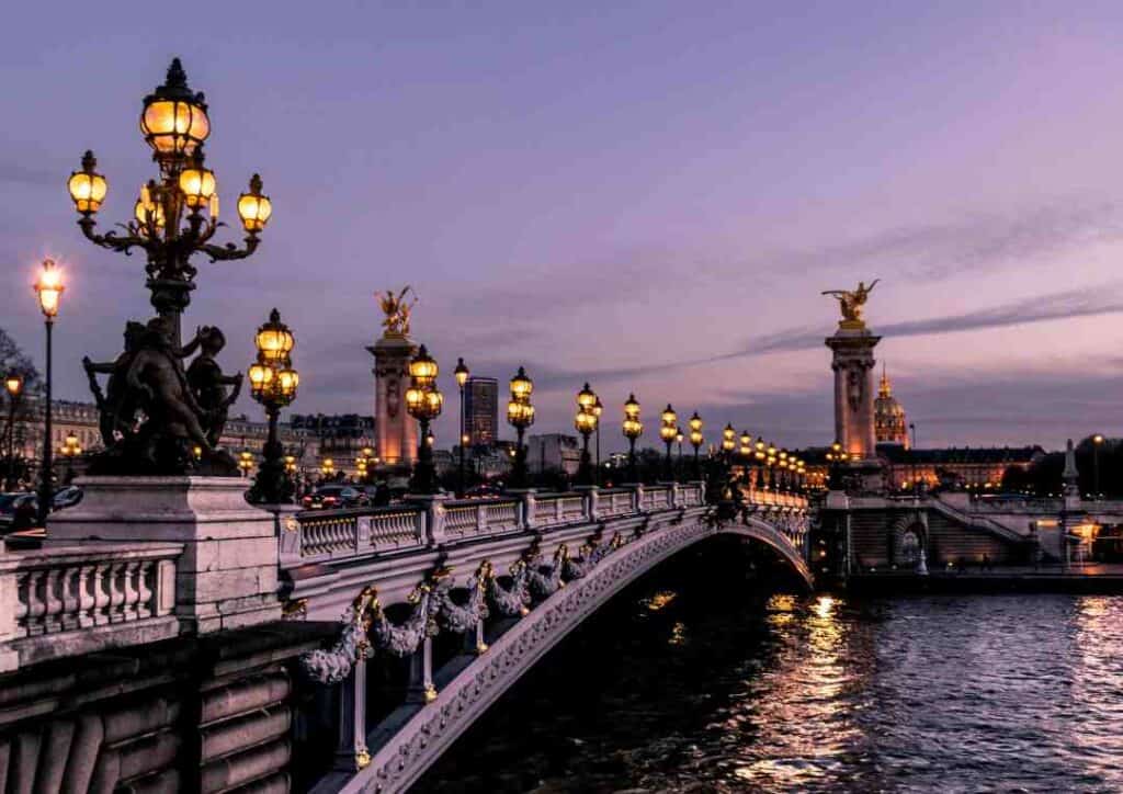 A beautiful old bridge with regal architecture in wihte looking silvery in the low winter light, decoreated with artsy datails, in the afternoon dusk with street lights providing a warm light over the surrounding elegant European city