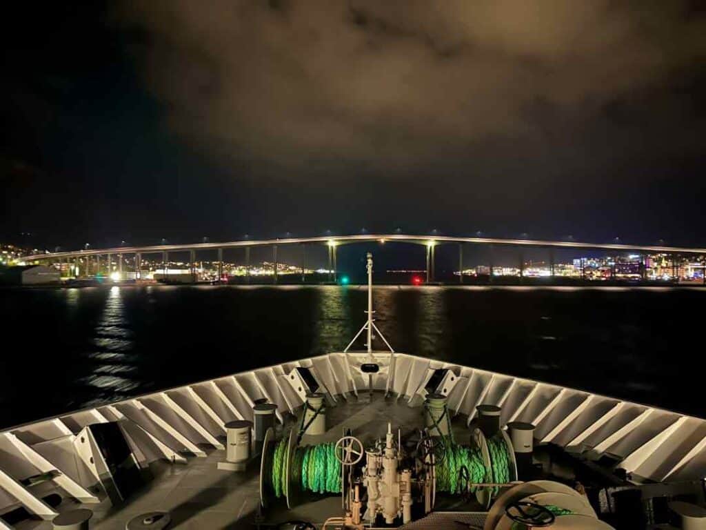 View from the white bau of a ship heading out a fjord at night, with a long bridge crossing ahead with streets light and cars and the city lights behind it, giving a golden glowing impression over the dark black water