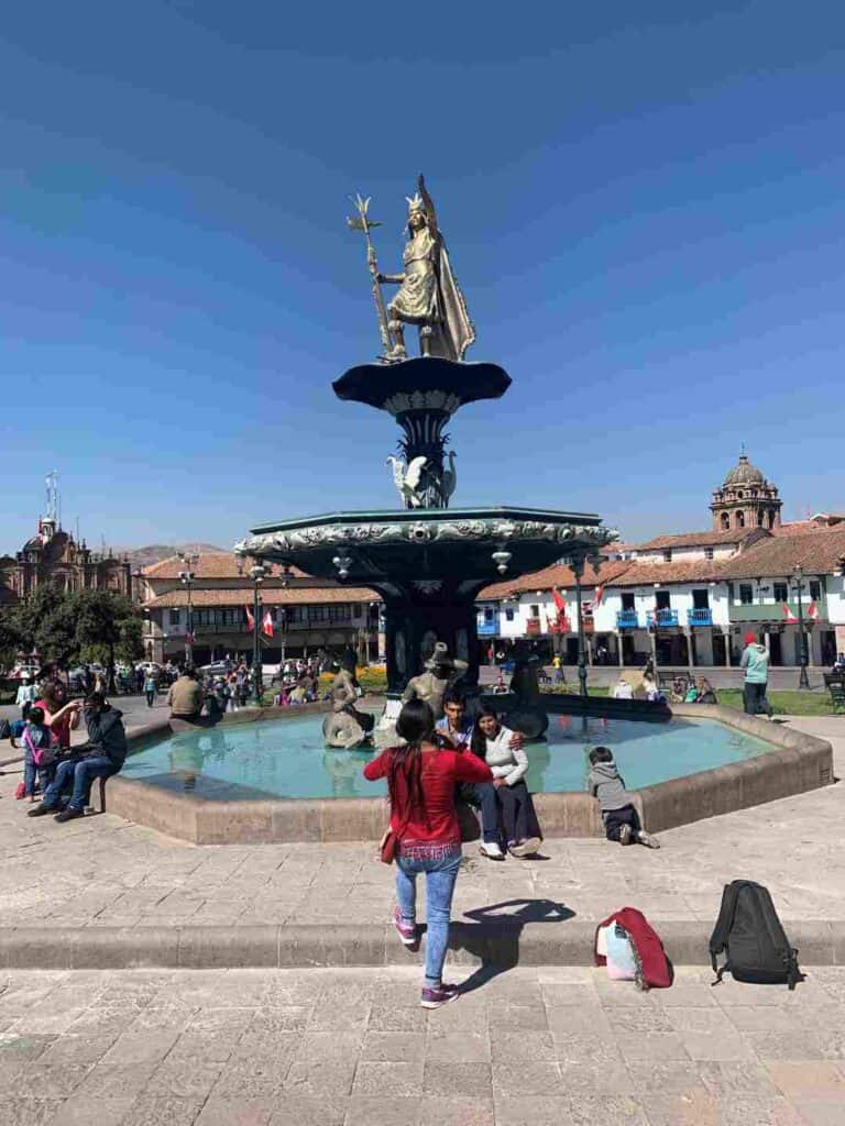 A bronze statue in the middle of a fountain on a town square surrounded by old white houses with terracotta roofs, and lots of people enjoying the plaza in the sun