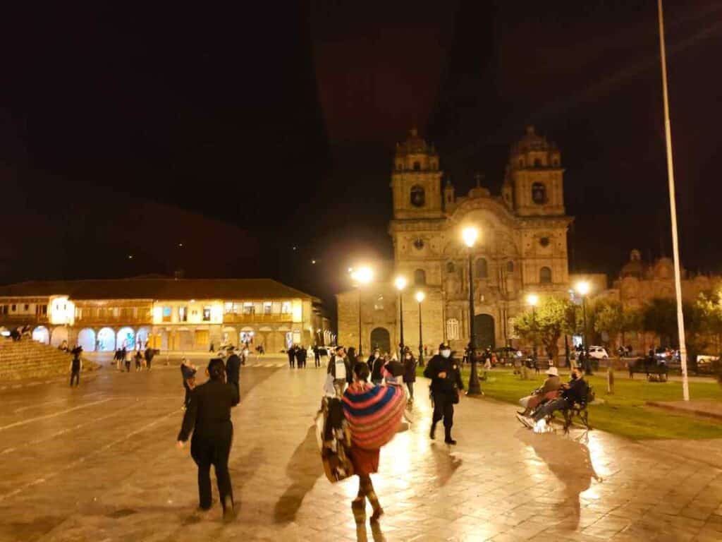 An old square with warm lighting at night with a beautiful stone church in the far and and people enjoying themselves in the squar