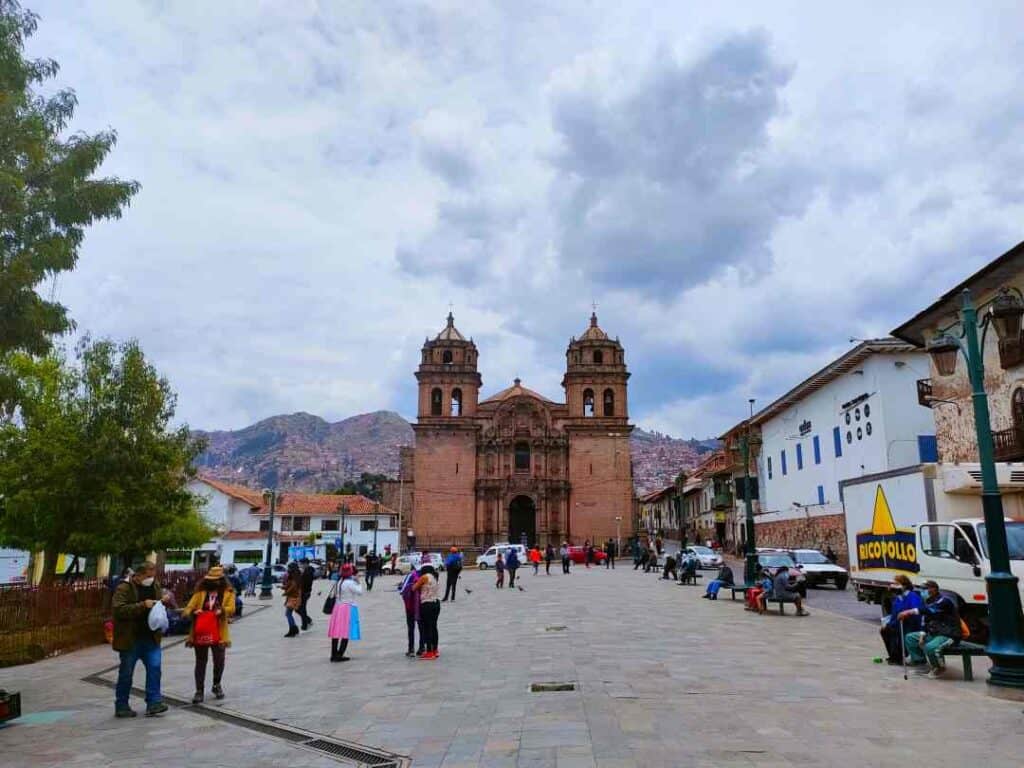 A wide town square with vibrant city life surrounded by white stone houses and a maroon stone cathedral, with mountains in the distance