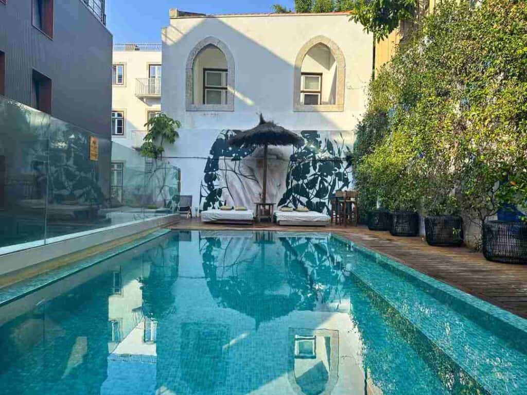 A blue pool in a charming backyard with green plants clinbing the walls, sun beds and classical buildings surrounding the pool area