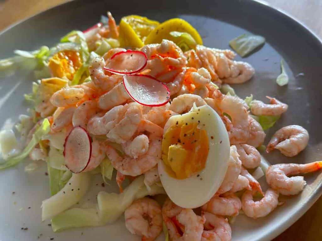 A huge scrimp sandwich up close, with pink shrimps, eggs, raddich and lemon on a dark plate