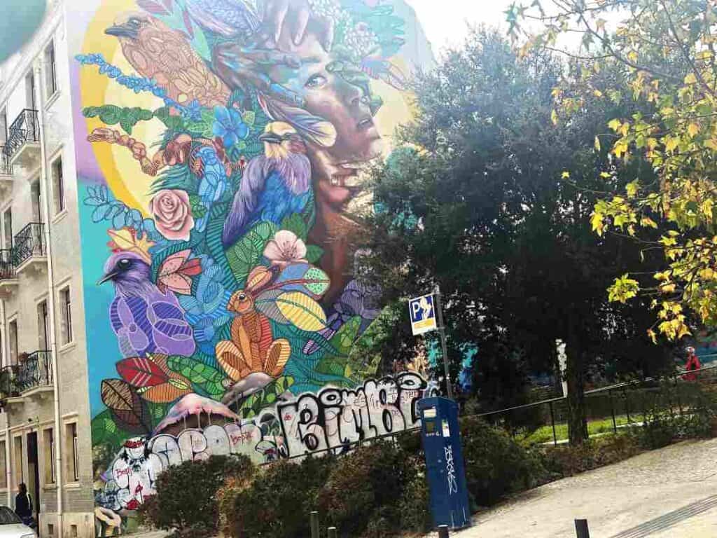 Colorful street art of nature, flowers and a human being on the entire side of a house wall behind large trees in a park