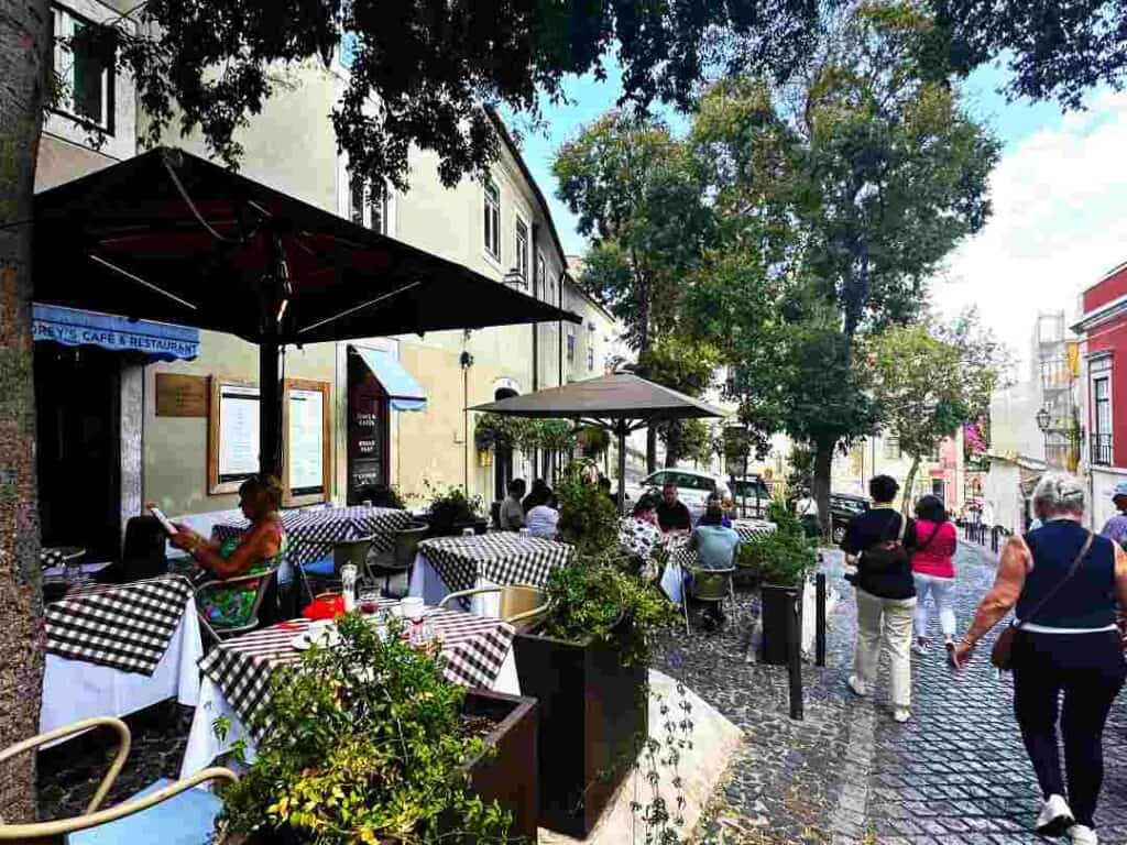 A street cafe next to narrow cobblestoned streets, with white and plaid table clothes, surrounded by colonial houses, trees, and greenery