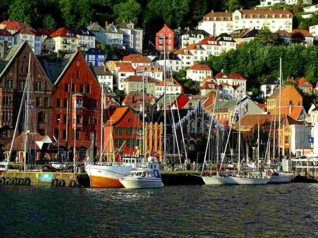 A harbor with dark blue water in front of a wharf with boats and old colorful charming buildings and white wooden houses in the green hills behind it on a sunny day