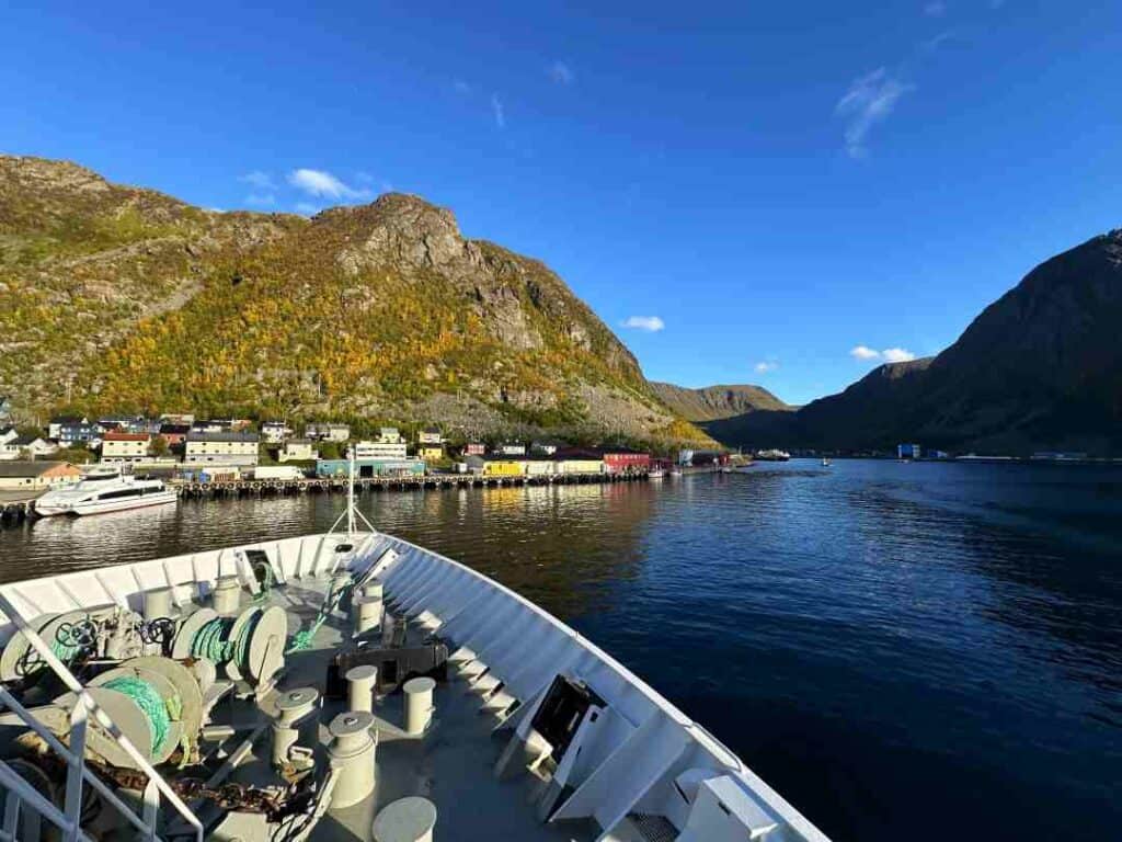 A white ship arriving at a small fishing village on a sunny summer day, with a green and barren Arctic mountain behind the village