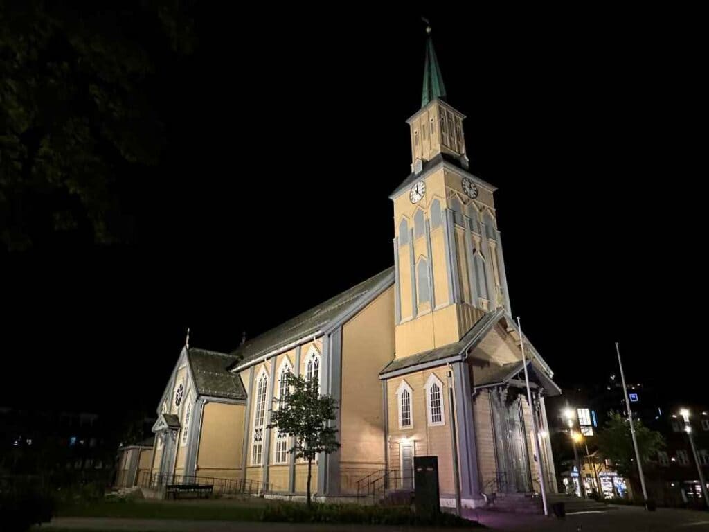 A pale yellow wooden church with grey elegant details and decorations at night, against the dark sky, lit by dozens of lights