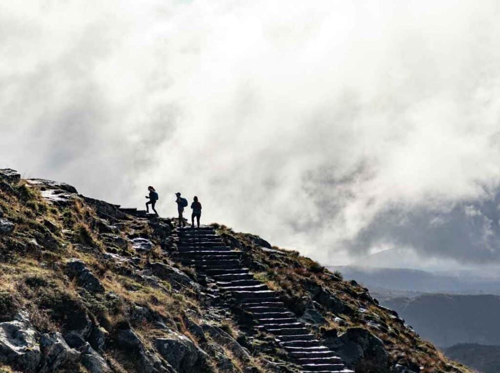 Three people hiking up a stairway on a mountain, with floating white clouds passing by and a whiff of sunshine sifting through