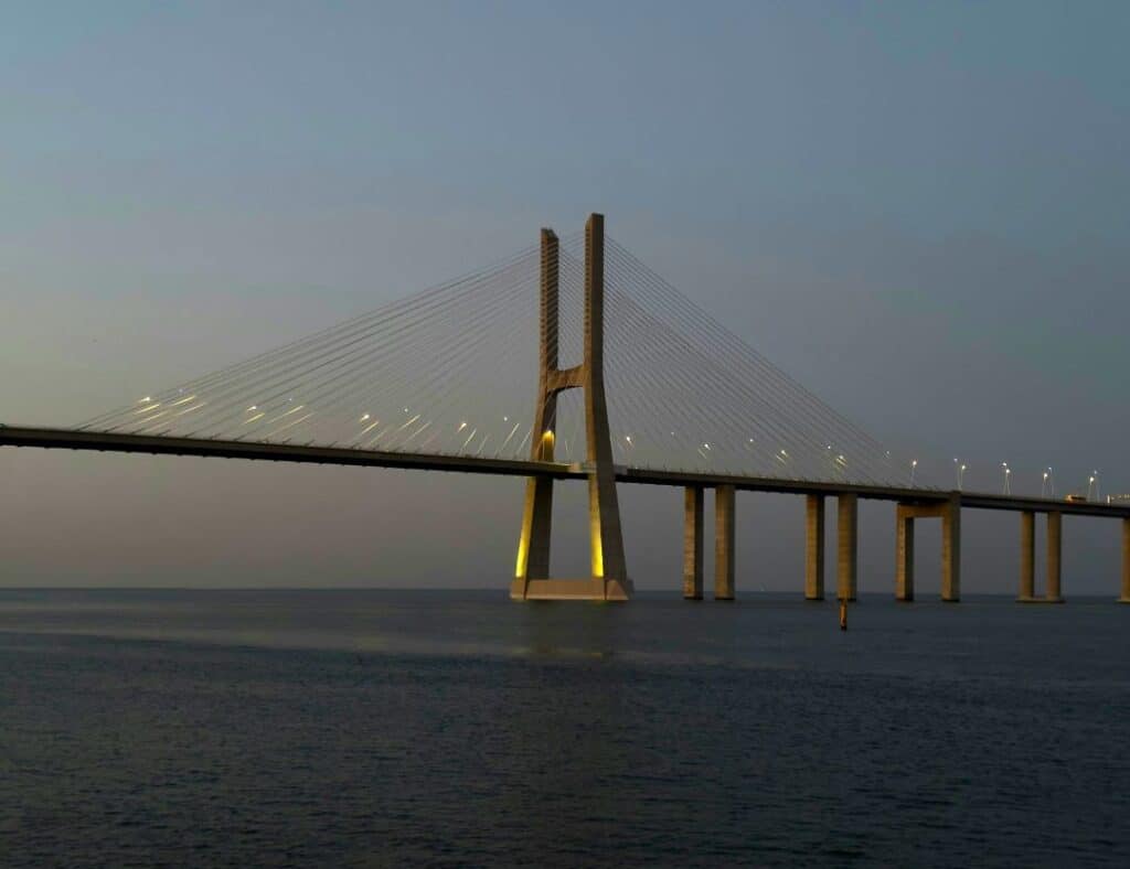 An impressive bridge structrue over dark blue water in golden colors at sunset, with thick columns supporting it into the water, and a fan shaped structure above making it a spectacular view