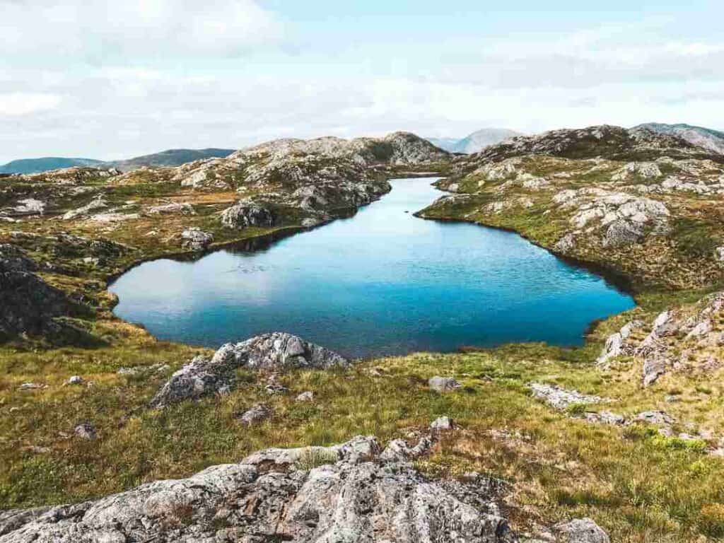 A mountain lake with still water reflecting the blue sky, surrounded by rocky mountain plains and greenery