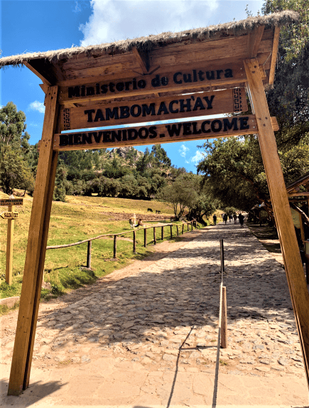 A wooden entry sign created as a portal, with a cobblestoned path starting inside heading into nature surrounded by big grees and grassy fields