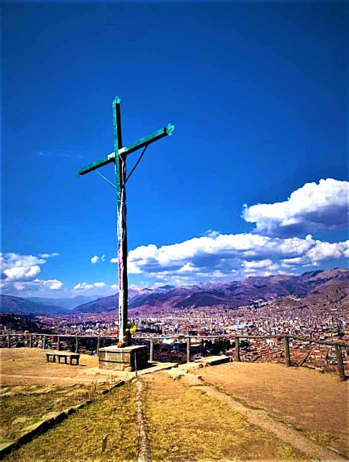 A large wooden cross towering high above a mountain city in the mountains below the viewpoint under blue skies with dotted white clouds