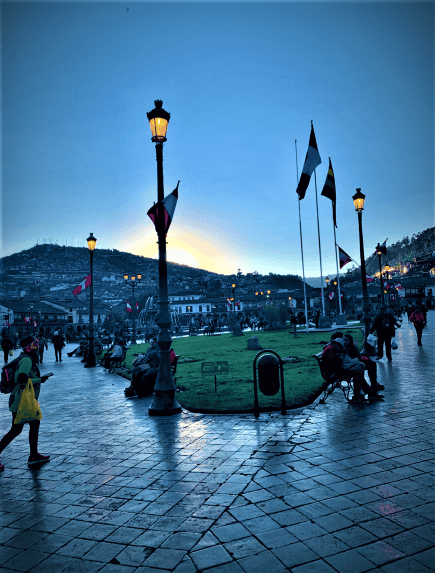 A park at dusk with lots of people still, flags on poles throughout the park, which is surrounded by hills under a pale blue sky