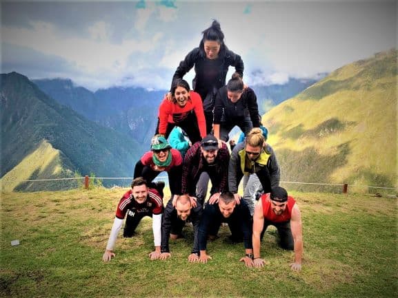 A group of travelers creating a human pyramide on a small plain high up in the mountains with stunning infinite mountain views in the background