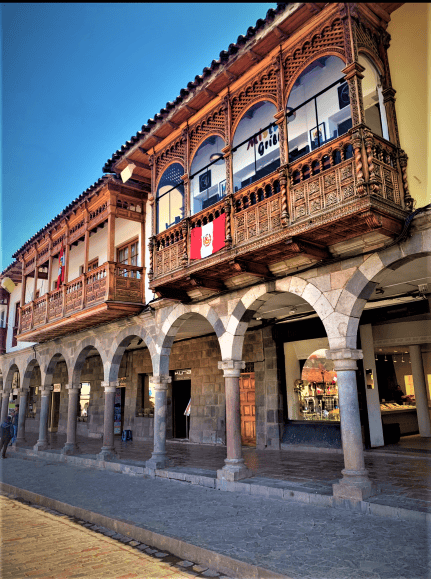 shop windows inside stone arches and columns, with an intricatly elaborate wooden balcony over in the second floor on a sunny day with blue skies above