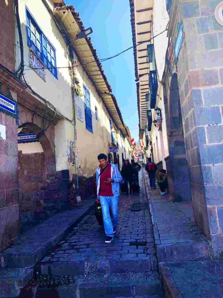 People walking in a narrow old town street on a sunny day, with cobblestones and brick houses in greys and colored details on both sides