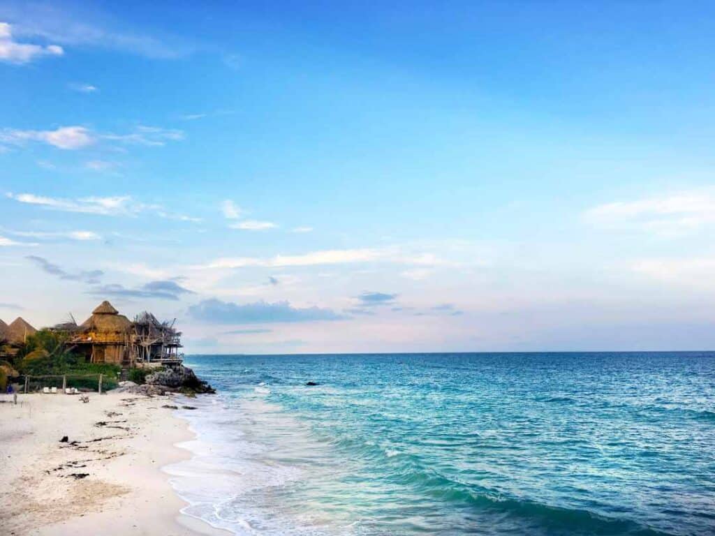 White sandy beach next to the deep blue water under a blue summer sky with wooden beach huts in the distance