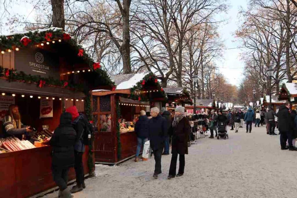 people strolling a Christmas market on a cold snowy day under naked trees in crisp air, along stalls selling christmas food and gifts