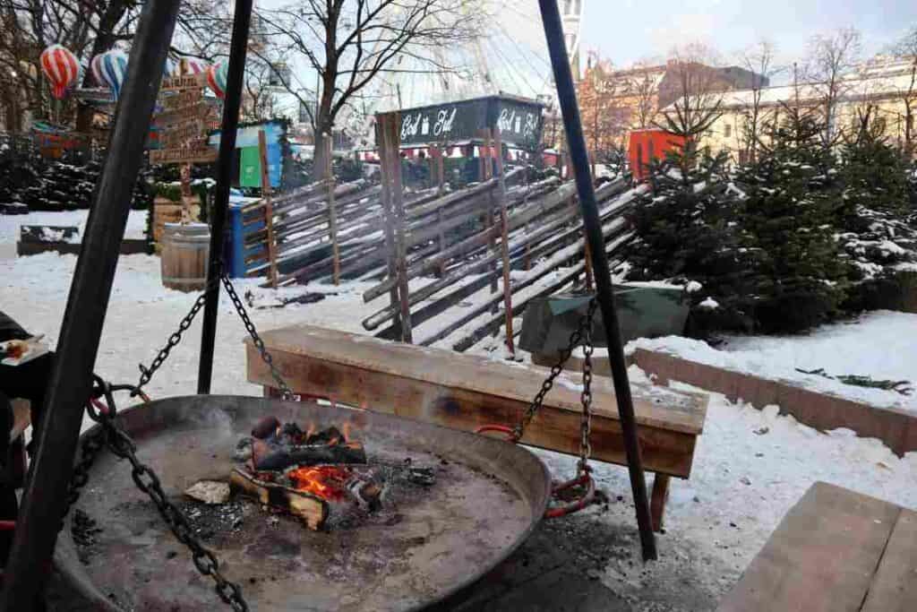 An outdoor bonfire next to wooden fences and christmas trees on a clear winter day with a pale blue sky above 