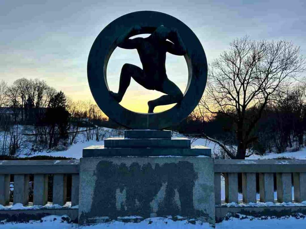 A sculpture of a man in a circle against a backdrop of a pale blue winter sky with the low sun setting behind naked trees and snowy forests