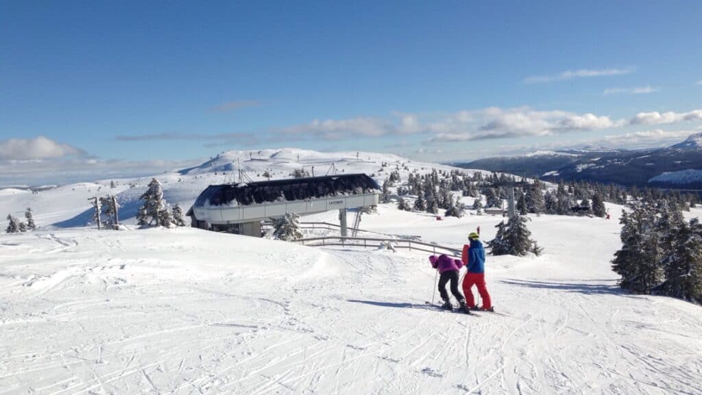 Two skiers standing on the top of a ski lift on white powdery snow on an alpine track under blue winter skies ready to go downhill